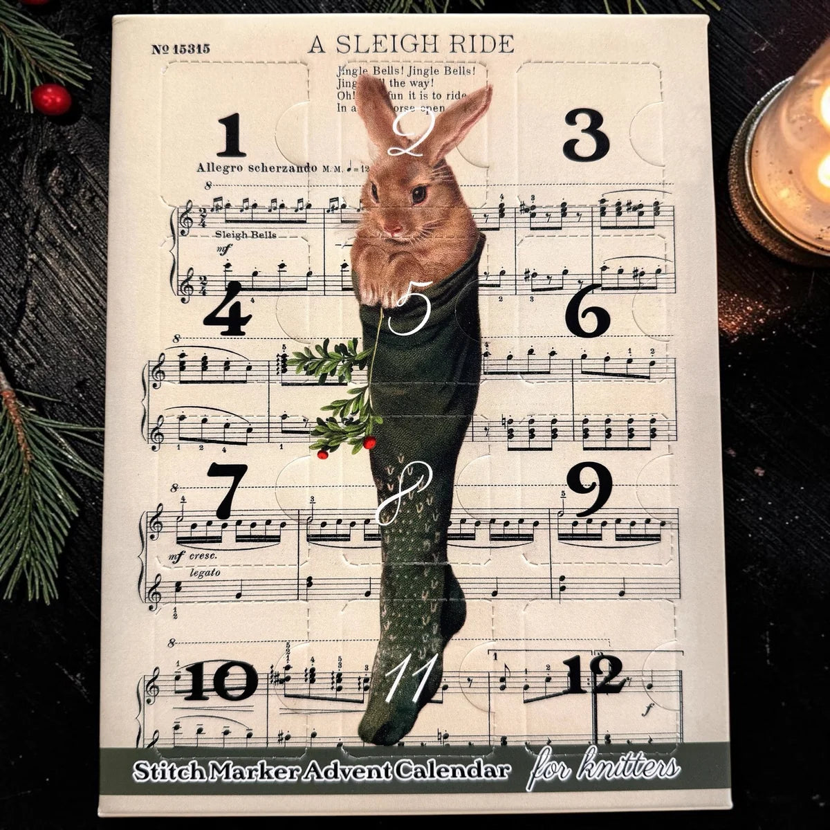 Firefly Notes Maschenmarkierer-Adventskalender 2026 für Strickende mit 12 Türchen, Vorderseite mit Hase im grünen Strickstrumpf vor Noten von „A Sleigh Ride“