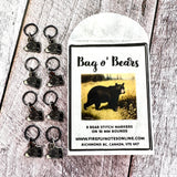 Firefly Notes Maschenmarkierer-Set Bag o' Bears aus Metall und Emaille, acht schwarze Bärenköpfe an schwarzen 10-mm-Ringen neben Papiertüte mit Vintage-Illustration eines Schwarzbären