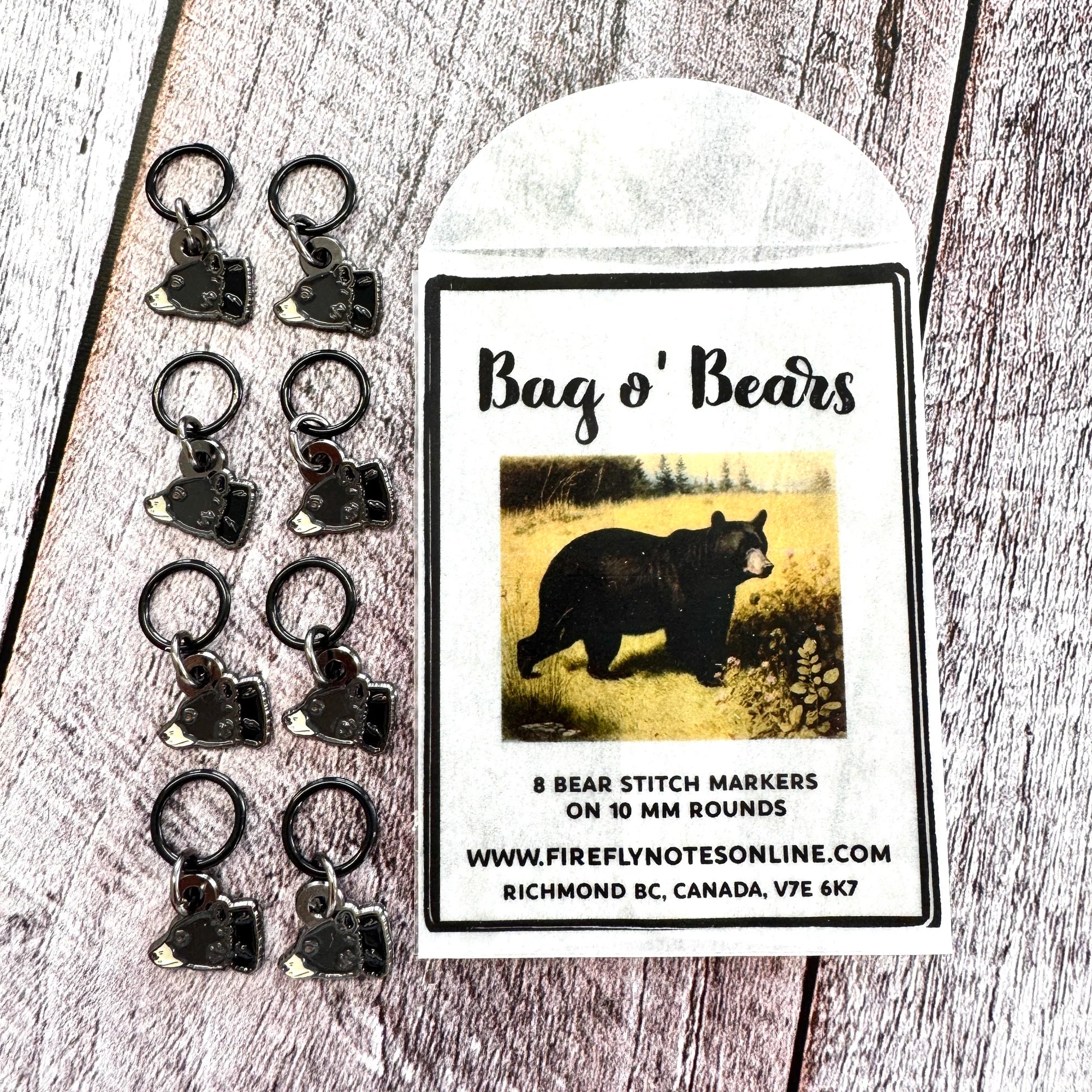 Firefly Notes Maschenmarkierer-Set Bag o' Bears aus Metall und Emaille, acht schwarze Bärenköpfe an schwarzen 10-mm-Ringen neben Papiertüte mit Vintage-Illustration eines Schwarzbären