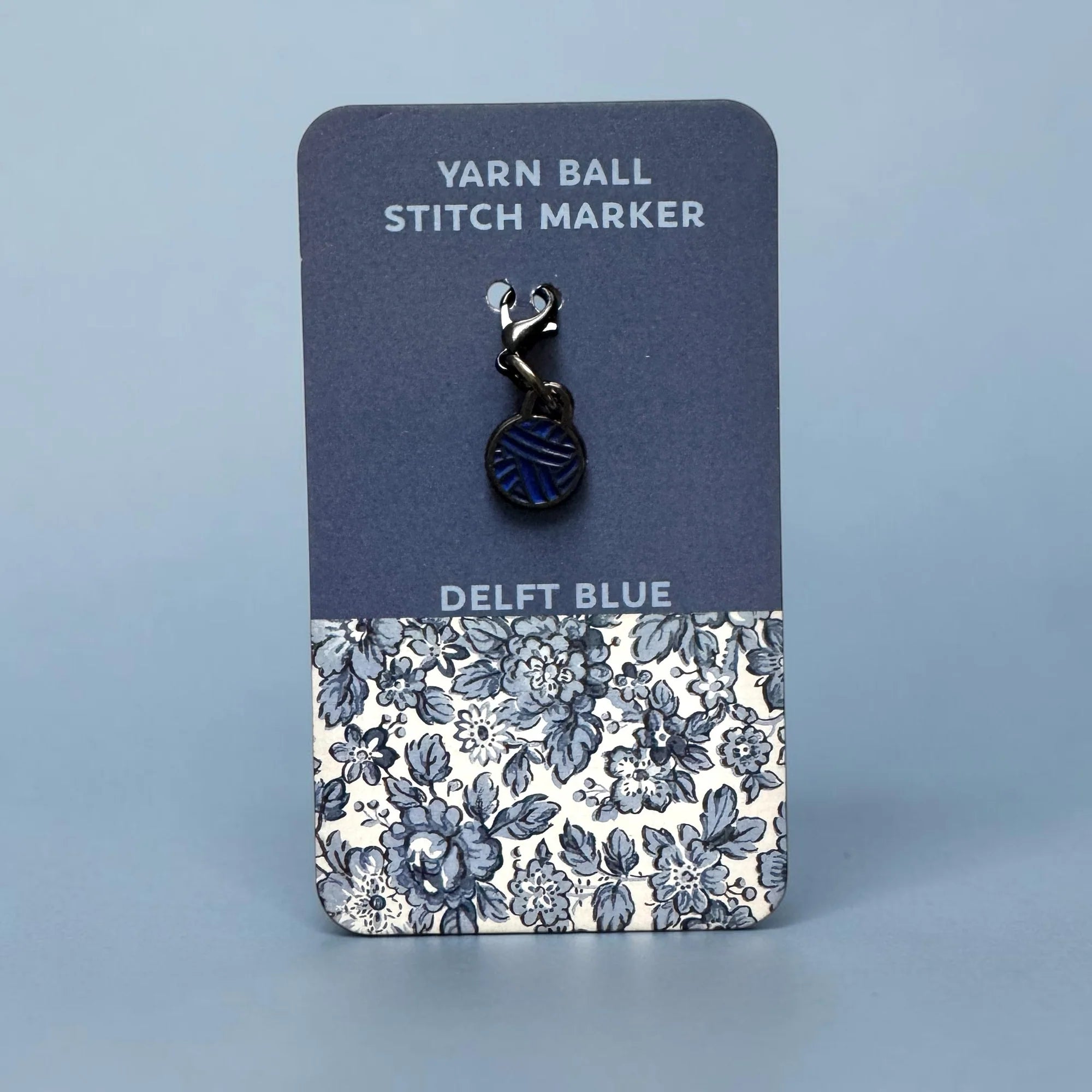 Firefly Notes Maschenmarkierer Yarn Ball in Delft Blue aus Metall und Emaille, blaues Wollknäuel mit schwarzem Hakenverschluss auf dunkelblauer Karte mit blau-weißem Blumenmuster